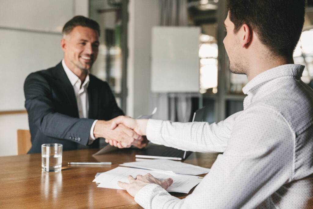 photo : Business, career and placement concept - successful young man smiling, and handshaking with european businessman after successful negotiations or interview in office