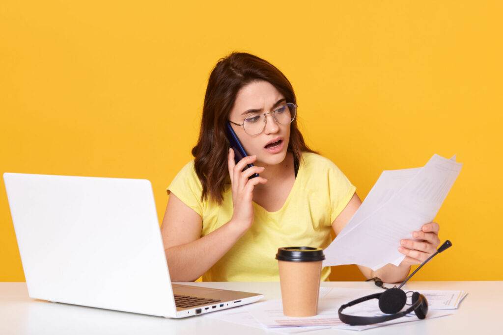 photo : Close up portrait of business woman working online via laptop, isolated over yellow studio background, talking to somebody via smart phone, holding paper in hands, looks astonished. and concentrated.