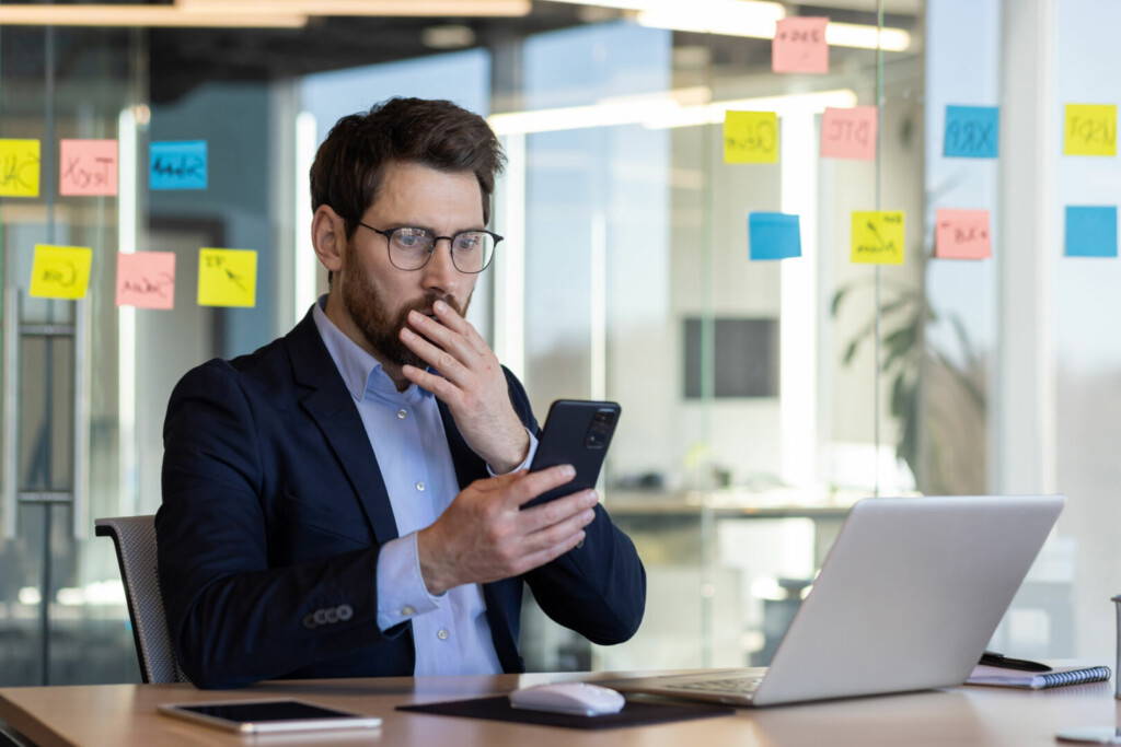 photo : Sad disappointed businessman reading news on phone, man in business suit using app on smartphone, looking upset, received bad news online in email, working with laptop inside office