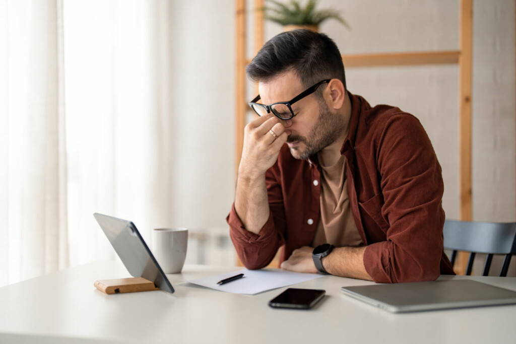 photo : Tired exhausted businessman rubbing eyes sitting in home office with portable technology devices on desk. Overworked burnout male manager with glasses feeling eyestrain pain after long work.