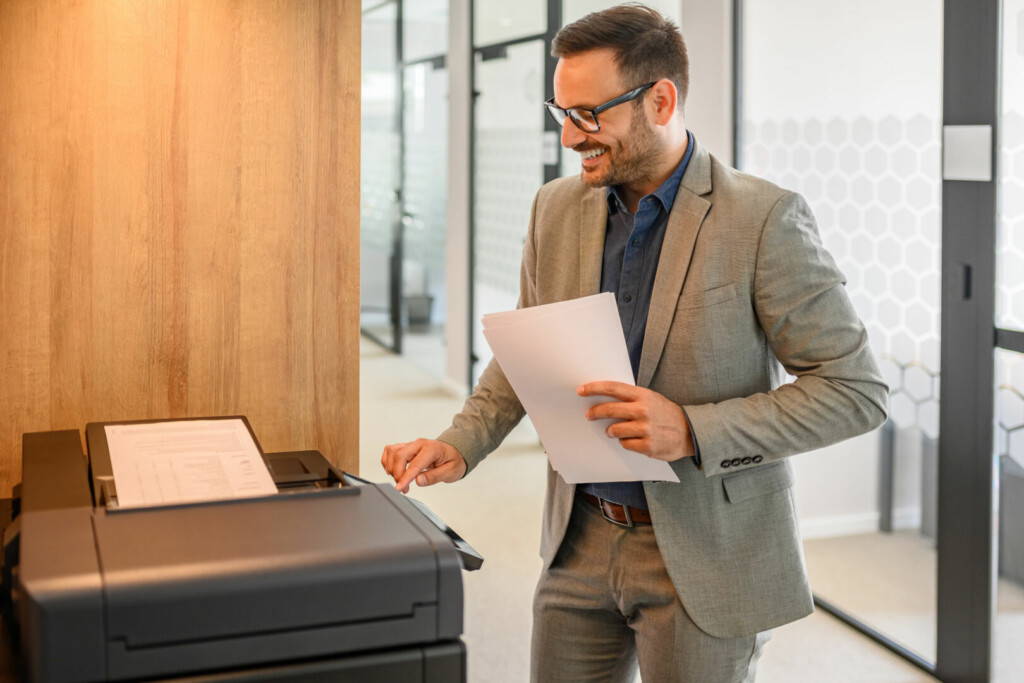 photo : Male smiling assistant in eyeglasses holding documents and operating modern computer printer in office