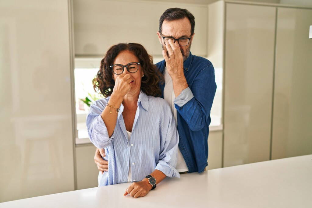 photo : Middle age couple standing together smelling something stinky and disgusting, intolerable smell, holding breath with fingers on nose. bad smell