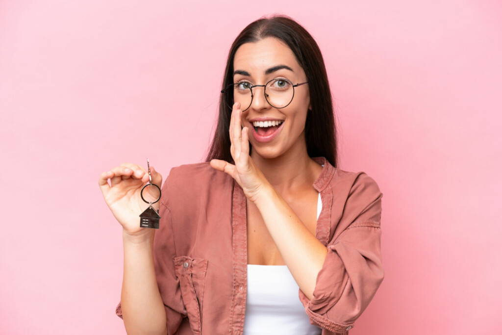 photo : Young woman holding home keys isolated on pink background whispering something