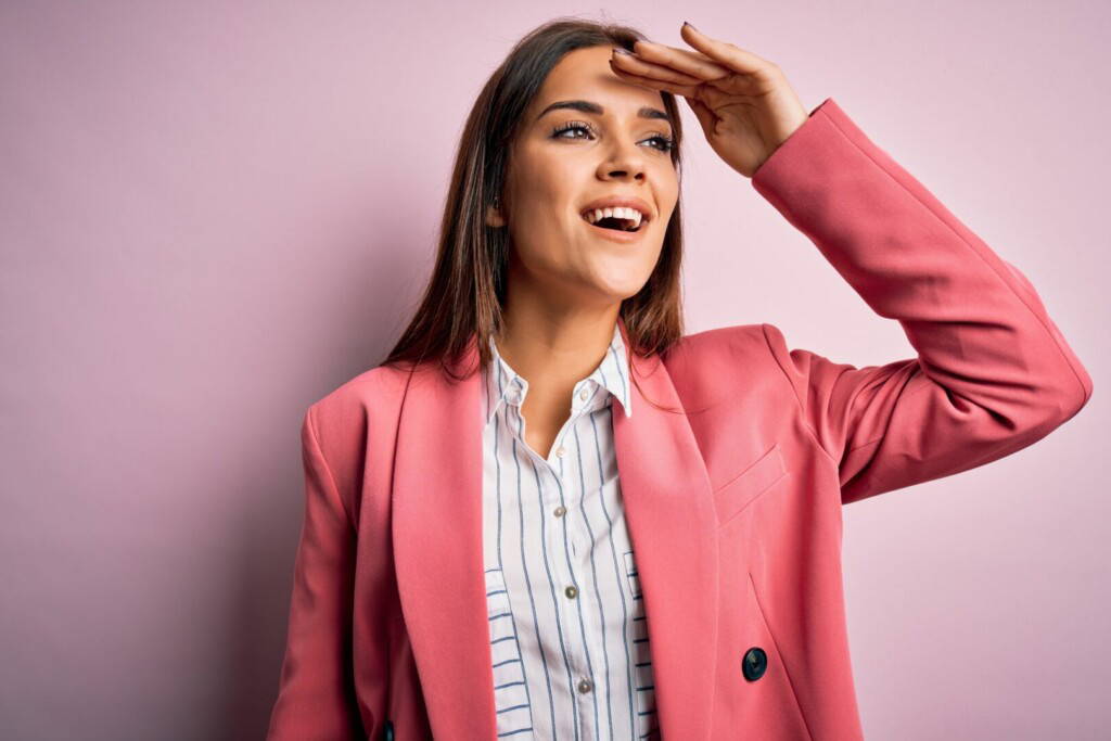 photo : Young beautiful brunette woman wearing jacket standing over isolated pink background very happy and smiling looking far away with hand over head. Searching concept.