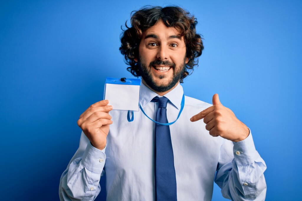 photo : Young handsome business man with beard holding id card identification over blue background with surprise face pointing finger to himself