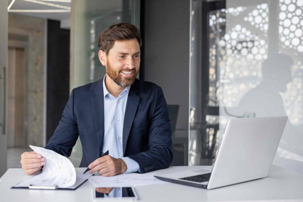 photo : Smiling young man working in office with documents, sitting at desktop with laptop and holding online meeting with clients and partners, signing agreement and invoices