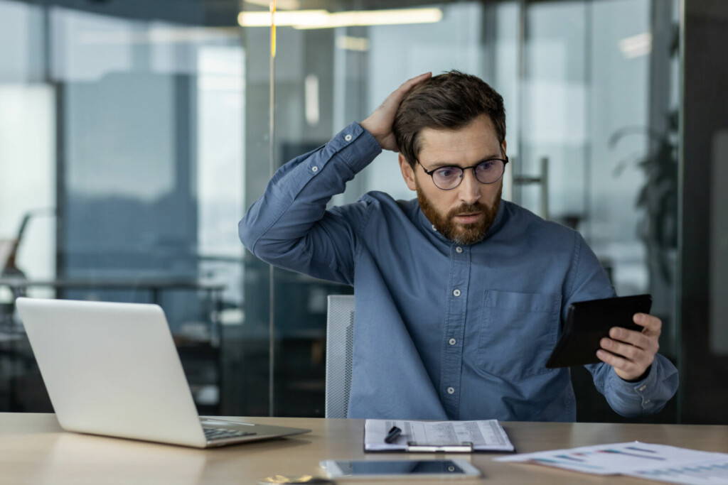 photo : Financial problems with accounts. Young bored man sitting in the office at the desk, working with documents and calculator, holding his head with his hand in frustration