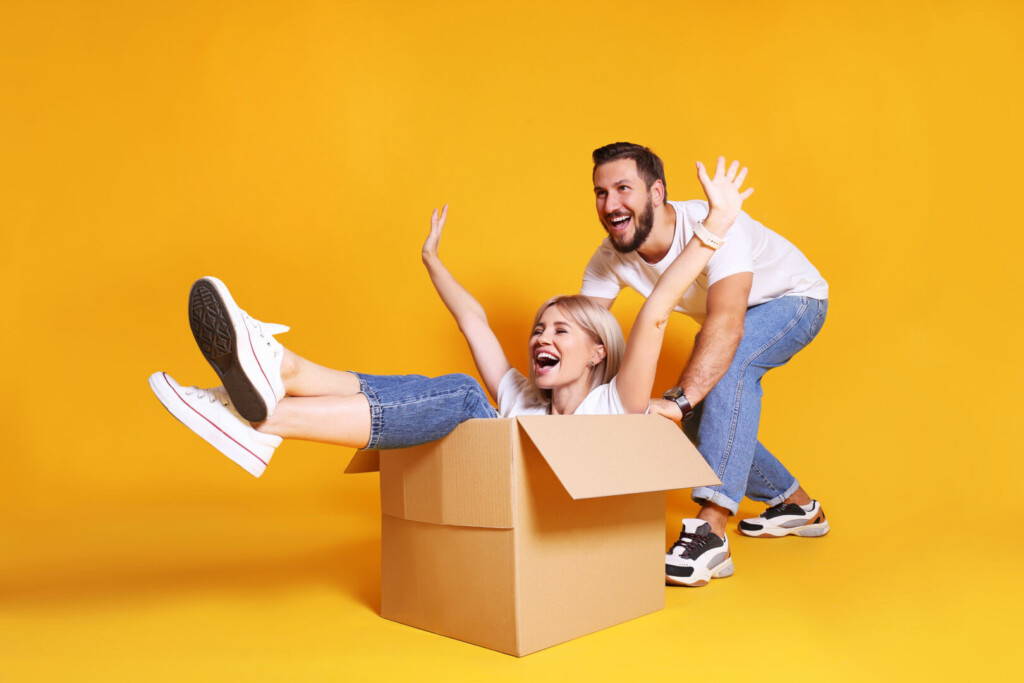 photo : Young married couple moving into a new home. Attractive blonde woman sitting in cardboard box while bearded man pushes her. Newely weds fooling around. Isolated yellow background, copy space, close up