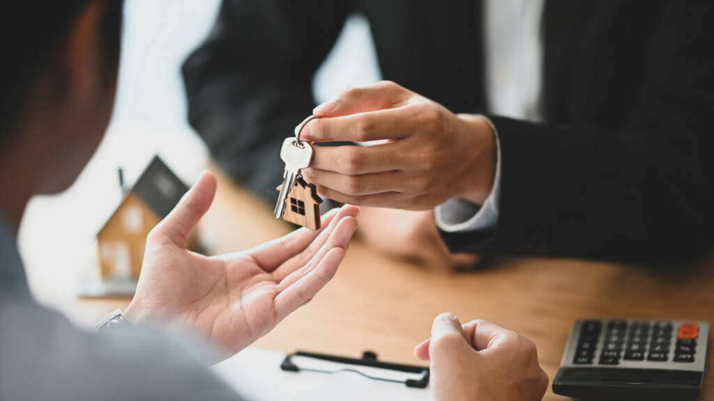 photo : Cropped shot of House broker giving a property key to his customer while he has writing/signing on agreement at the modern wooden desk. Loan, Debt, Credit, Buying or Selling agreement concept.